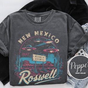 May include: A dark gray t-shirt with a vintage graphic design featuring a UFO and the text "New Mexico Roswell 1947" and "Welcome to Roswell".
