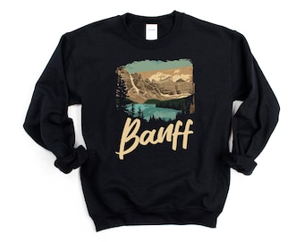 Puede incluir: Sudadera negra con un estampado gráfico de una cordillera y un lago. El texto "Banff" está impreso en color crema debajo del gráfico.
