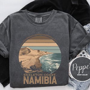 Peut inclure: Un t-shirt gris avec un graphique de style vintage d'une côte désertique avec le texte "Skeleton Coast, Namibia".