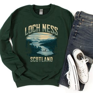 May include: A dark green sweatshirt with a vintage-style graphic of Loch Ness, Scotland. The graphic features a lake with a silhouette of the Loch Ness Monster and the text "Loch Ness Scotland".