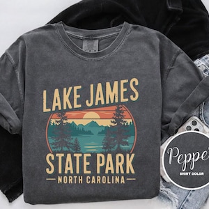 A gray t-shirt with a graphic design featuring a sunset over a lake and mountains, along with the text "Lake James State Park North Carolina".