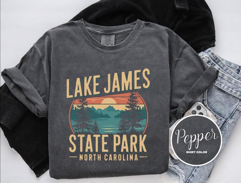 A gray t-shirt with a graphic design featuring a sunset over a lake and mountains, along with the text "Lake James State Park North Carolina".