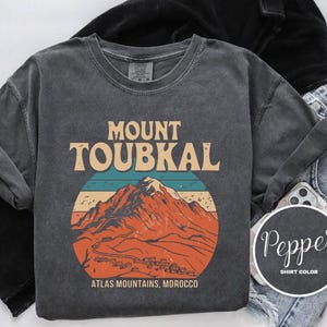 May include: A dark grey t-shirt with a vintage-style graphic of Mount Toubkal in the Atlas Mountains of Morocco. The text "Mount Toubkal" is in a bold, retro font. The shirt has a distressed look and feel.