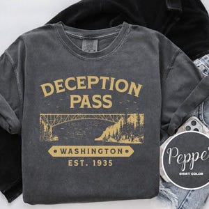 Puede incluir: Una camiseta gris oscuro con un gráfico de estilo vintage de un puente y el texto "Deception Pass, Washington Est. 1935".