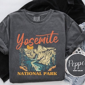 Puede incluir: Una camiseta gris oscuro con un gráfico de estilo vintage del Parque Nacional de Yosemite. El gráfico presenta una cordillera, un río y una puesta de sol. El texto "Yosemite National Park" está impreso debajo del gráfico.