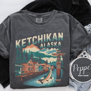 以下が含まれることがあります： 暗いグレーのTシャツに、ヴィンテージスタイルの街並みを山とトーテムポールで描いたグラフィックがプリントされています。Tシャツには「Ketchikan Alaska」という文字が白でプリントされています。