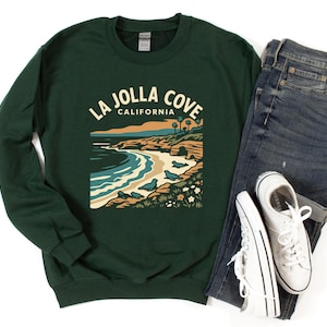 May include: Dark green sweatshirt with a graphic of La Jolla Cove, California. The design includes the text "La Jolla Cove California", a beach scene, and seals. The sweatshirt is paired with blue jeans and white sneakers.