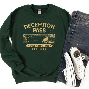 May include: A dark green sweatshirt with the text "Deception Pass" in gold lettering. The sweatshirt also features a graphic of a bridge and the text "Washington Est. 1935".
