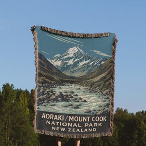 May include: A teal and beige woven blanket depicting a landscape of Aoraki/Mount Cook National Park in New Zealand. The blanket shows a mountain range, river, and text that reads "AORAKI/MOUNT COOK NATIONAL PARK NEW ZEALAND". The blanket has a fringed edge.