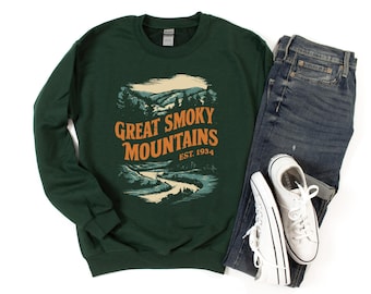 Könnte beinhalten: Ein dunkelgrüner Pullover mit einem Vintage-Grafikdruck eines Gebirges und eines Flusses. Der Text "Great Smoky Mountains Est. 1934" ist auf der Vorderseite des Pullovers aufgedruckt.