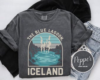 May include: A dark gray t-shirt with a vintage-style graphic of the Blue Lagoon in Iceland. The graphic features a blue lagoon with steam rising from it, surrounded by rocks and mountains. The text "The Blue Lagoon, Iceland" is printed below the graphic.