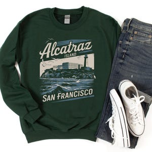 May include: A dark green sweatshirt with a vintage-style graphic of Alcatraz Island, San Francisco. The graphic features a lighthouse, buildings, and the text "Alcatraz Island, San Francisco".