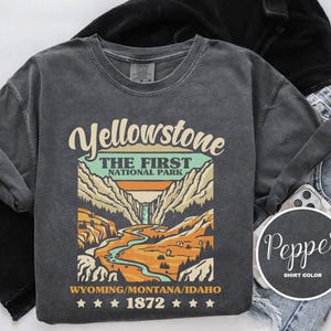 May include: A dark gray t-shirt with a vintage-style graphic of Yellowstone National Park. The graphic features a mountain range, a river, and the text "Yellowstone, The First National Park, Wyoming/Montana/Idaho, 1872".