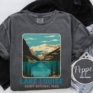 May include: Dark gray sweatshirt featuring a scenic illustration of Lake Louise in Banff National Park. The design includes a lake, mountains, and trees, with the text "LAKE LOUISE BANFF NATIONAL PARK" in teal. The sweatshirt is paired with black clothing and denim jeans.