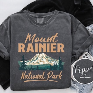 May include: A dark gray t-shirt with a vintage look featuring a graphic of Mount Rainier National Park. The text "Mount Rainier National Park" is printed in brown on the shirt.