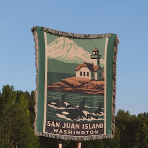 May include: A green and beige woven throw blanket featuring a scenic illustration of San Juan Island, Washington. The design includes a lighthouse, orcas, and a mountain. The blanket has a fringed edge.