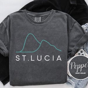 May include: A dark gray, long-sleeved t-shirt with the text "ST. LUCIA" in white. Above the text is a teal-colored mountain graphic. The shirt is folded, with a black jacket and blue jeans visible in the background. A round sticker with the word "Pepper" is also visible.