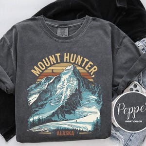 May include: Dark gray Mount Hunter Alaska graphic t-shirt. The design features a mountain range with a sunset backdrop and the text "Mount Hunter, Alaska". The shirt is displayed with black clothing and blue jeans.