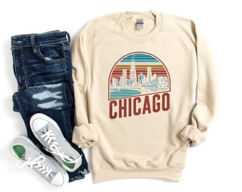 Puede incluir: Sudadera color crema con un diseño retro de la silueta de Chicago. El gráfico presenta un paisaje urbano, un puente, un tren y el nombre de la ciudad en negrita. Combinada con vaqueros azules y zapatillas grises.