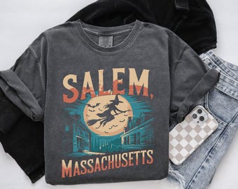 Könnte beinhalten: Ein dunkelgraues T-Shirt mit einer Grafik im Vintage-Stil einer Hexe, die auf einem Besen vor einem Mond und Häusern fliegt. Der Text "SALEM, MASSACHUSETTS" ist auf das Shirt gedruckt.