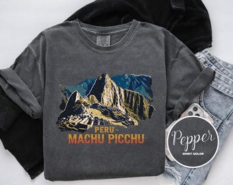 Puede incluir: Una camiseta gris oscura con un gráfico de Machu Picchu, una famosa ciudadela inca en Perú. El texto "Perú - Machu Picchu" está impreso debajo de la imagen.