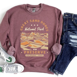 May include: A maroon sweatshirt with the text "Great Sand Dunes National Park and Preserve, Colorado - 2004" and a graphic of sand dunes. The sweatshirt is paired with blue jeans.