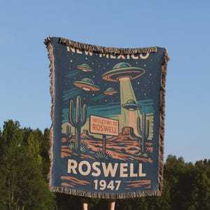 May include: A navy blue throw blanket featuring a retro Roswell, New Mexico design. The blanket displays flying saucers, cacti, and the text "Welcome to Roswell" and "1947". The blanket has a fringed edge.