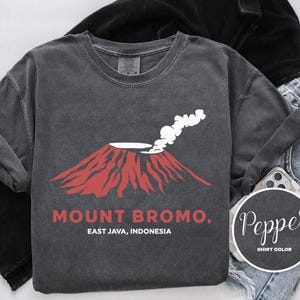 Könnte beinhalten: Dunkelgraues Langarmshirt mit roter Vulkan-Grafik und weißem Rauch, sowie dem Text "MOUNT BROMO. EAST JAVA, INDONESIA." Das Shirt wird mit blauen Jeans und einer schwarzen Jacke kombiniert. Ein Handy ist ebenfalls zu sehen.