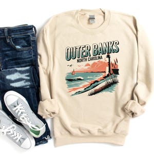 Può includere: Felpa color crema con grafica delle Outer Banks, Carolina del Nord, con faro, onde oceaniche e la scritta "Outer Banks North Carolina". La felpa è abbinata a jeans blu e scarpe da ginnastica grigie.