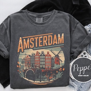 Może przedstawiać: Koszulka z długim rękawem w kolorze pieprzu z napisem "AMSTERDAM" w kolorze pomarańczowym i białym. Grafika przedstawia holenderski krajobraz miejski z budynkami, wiatrakiem i rowerem. Koszulka jest połączona z czarnymi ubraniami i dżinsami.