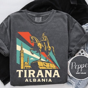 May include: Dark gray t-shirt featuring a graphic of a horse and rider statue, a mosque, and the text "TIRANA ALBANIA" in white. The design includes red, blue, and yellow accents. The shirt is displayed with black clothing and blue jeans.