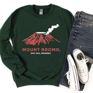 Op de afbeelding: Donkergroene sweatshirt met een rode en witte afbeelding van een vulkaan, met de tekst "MOUNT BROMO. EAST JAVA, INDONESIA". De sweatshirt wordt gecombineerd met een blauwe jeans en witte sneakers.
