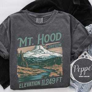 May include: A dark gray t-shirt with a vintage illustration of Mt. Hood, Oregon. The text "MT. HOOD OREGON" is printed above the illustration. The text "ELEVATION 11,249 FT" is printed below the illustration.