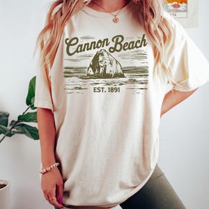 Peut inclure: Un t-shirt beige surdimensionné avec un imprimé graphique d'une formation rocheuse dans l'océan. Le graphique indique "Cannon Beach EST. 1891".