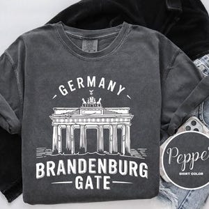 May include: Dark gray crew neck t-shirt with a white graphic of the Brandenburg Gate and the words "GERMANY" and "BRANDENBURG GATE". The shirt is folded with a black jacket and blue jeans.