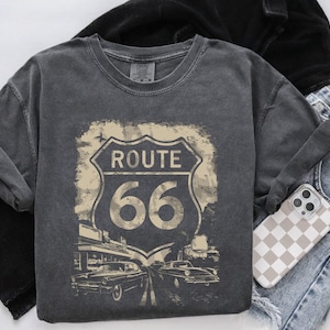 Peut inclure: Un t-shirt gris foncé avec un graphique de style vintage d'un panneau Route 66. Le panneau est blanc avec du texte noir et un numéro 66 rouge. Le graphique est entouré d'un fond délavé et vieilli.
