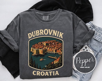 Op de afbeelding: Donkergrijze t-shirt met een afbeelding van Dubrovnik, Kroatië. Het ontwerp toont een illustratie van een kuststad met de tekst "DUBROVNIK" erboven en "CROATIA" eronder. Het shirt wordt getoond met opgerolde mouwen en een spijkerbroek.