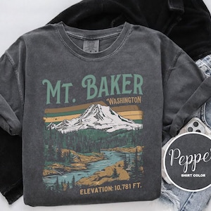 Może przedstawiać: Szary t-shirt z nadrukiem w stylu vintage przedstawiającym Mt. Baker w stanie Waszyngton. Grafika przedstawia pasmo g&oacute;rskie, drzewa i rzekę. Na koszulce nadrukowano tekst "Mt. Baker, Washington" i "Elevation: 10,781 ft."