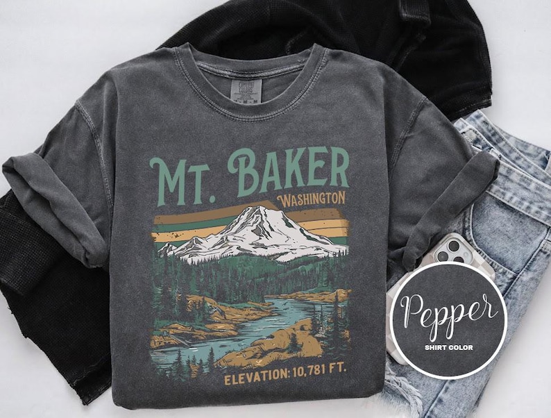 Może przedstawiać: Szary t-shirt z nadrukiem w stylu vintage przedstawiającym Mt. Baker w stanie Waszyngton. Grafika przedstawia pasmo g&oacute;rskie, drzewa i rzekę. Na koszulce nadrukowano tekst "Mt. Baker, Washington" i "Elevation: 10,781 ft."
