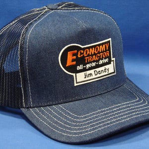 Może przedstawiać: Niebieska czapka typu trucker z dżinsu z siatkowym tyłem. Czapka posiada haftowaną naszywkę z napisem "ECONOMY TRACTOR all-gear-drive Jim Dandy" w kolorze pomarańczowym i białym. Białe przeszycia zdobią daszek.