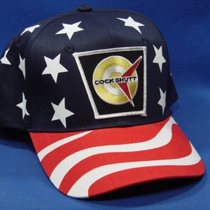 May include: A blue baseball cap with white stars and a red, white, and blue wave pattern on the brim. The cap has a patch with the words "Cock Shutt" in gold lettering.