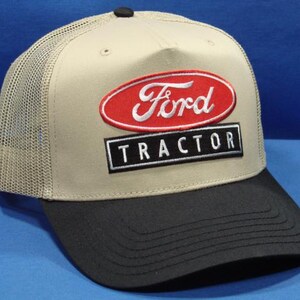 Könnte beinhalten: Eine beige-schwarze Trucker-Cap mit Netzrückseite und festem Vorderteil. Auf der Vorderseite befindet sich ein gestickter roter ovaler Aufnäher mit dem weißen Schriftzug Ford und darunter ein schwarzer rechteckiger Aufnäher mit dem weißen Text TRACTOR.