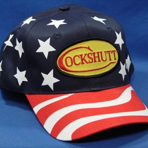 May include: A blue baseball cap with a red, white, and blue American flag design on the brim. The front of the cap features a white star pattern and a yellow oval patch with the text "COCKSHUTT".
