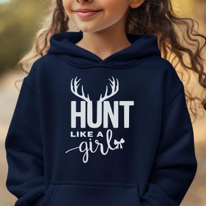 May include: A navy blue hooded sweatshirt featuring a white graphic print on the chest. The design includes a pair of deer antlers above the text HUNT LIKE A girl, with the word girl written in a cursive script and ending with a small bow icon.