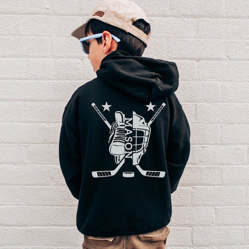 Puede incluir: Sudadera con capucha negra con un dise&ntilde;o de hockey blanco en la espalda. El dise&ntilde;o incluye un pat&iacute;n de hockey, casco, palos cruzados, un disco y el nombre "MASON". Dos estrellas est&aacute;n encima de los palos.