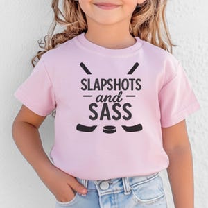 Op de afbeelding: Een lichtroze t-shirt met de zwarte tekst "SLAPSHOTS and SASS", samen met afbeeldingen van hockeysticks en een puck. Het shirt met korte mouwen lijkt van een zacht materiaal te zijn.