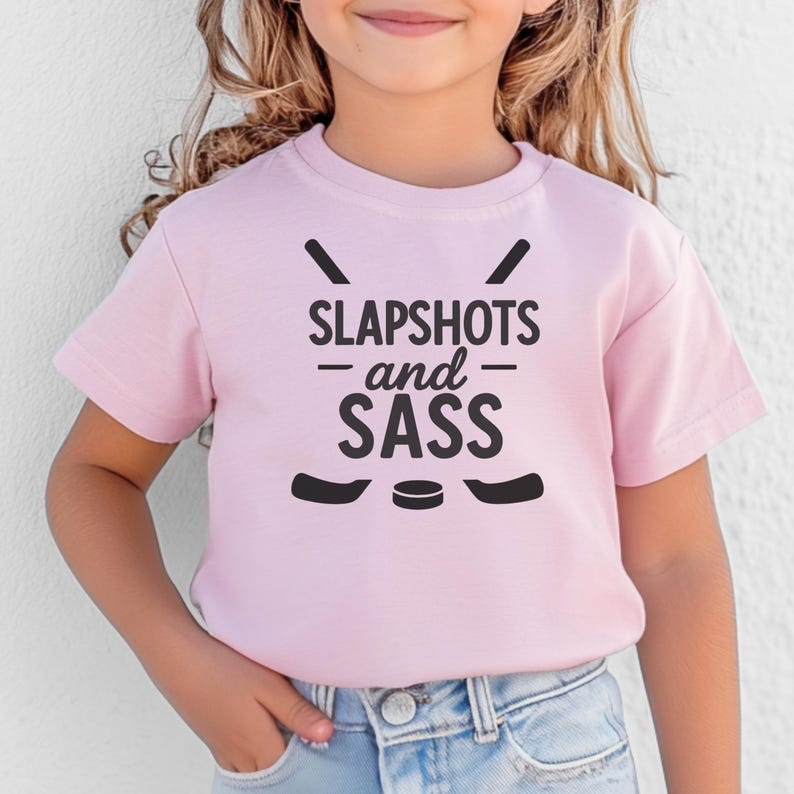 Op de afbeelding: Een lichtroze t-shirt met de zwarte tekst "SLAPSHOTS and SASS", samen met afbeeldingen van hockeysticks en een puck. Het shirt met korte mouwen lijkt van een zacht materiaal te zijn.