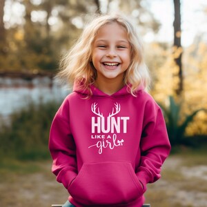 May include: A bright magenta hooded sweatshirt featuring a white graphic print on the chest. The design includes a pair of deer antlers above the text HUNT LIKE A GIRL, with a small bow icon integrated into the lettering of the word girl.