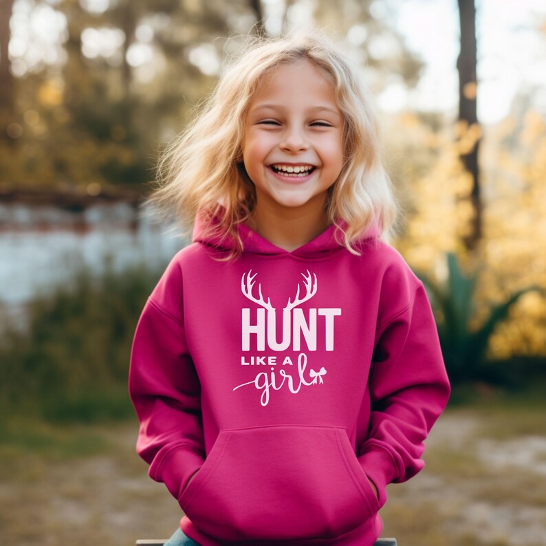 May include: A bright magenta hooded sweatshirt featuring a white graphic print on the chest. The design includes a pair of deer antlers above the text HUNT LIKE A GIRL, with a small bow icon integrated into the lettering of the word girl.