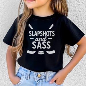 Op de afbeelding: Zwart T-shirt met de witte tekst "SLAPSHOTS and SASS". Het ontwerp bevat hockeysticks en een puck. Het shirt wordt door een kind gedragen.